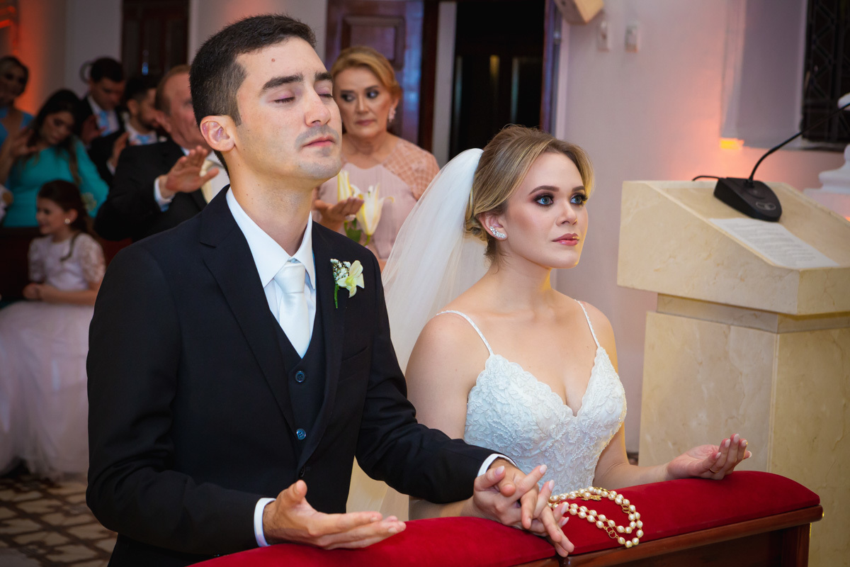 Celebração do casamento na Igreja Santa Terezinha, noivos ajoelhados recebendo benção do padre.