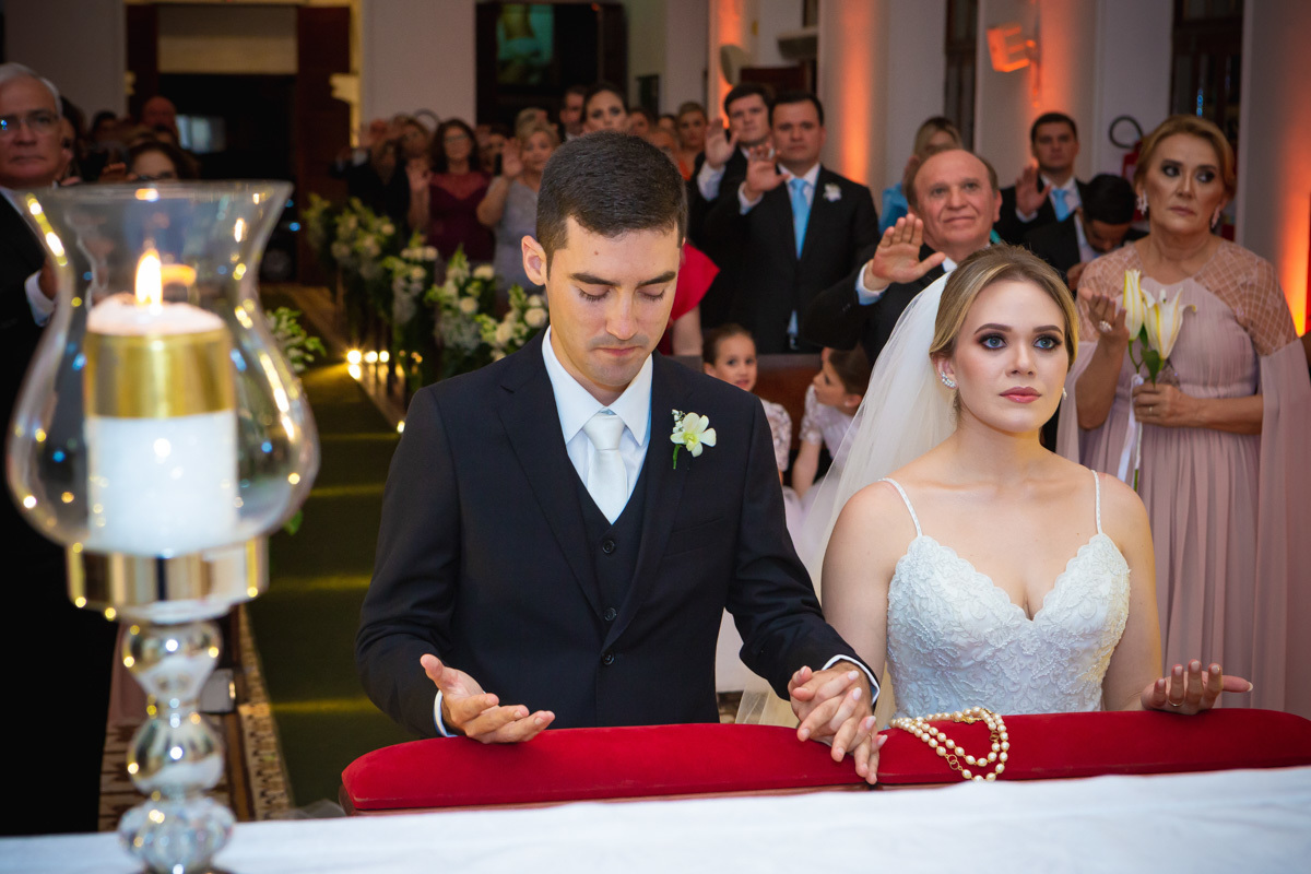 Celebração do casamento na Igreja Santa Terezinha, noivos ajoelhados de mãos dadas.