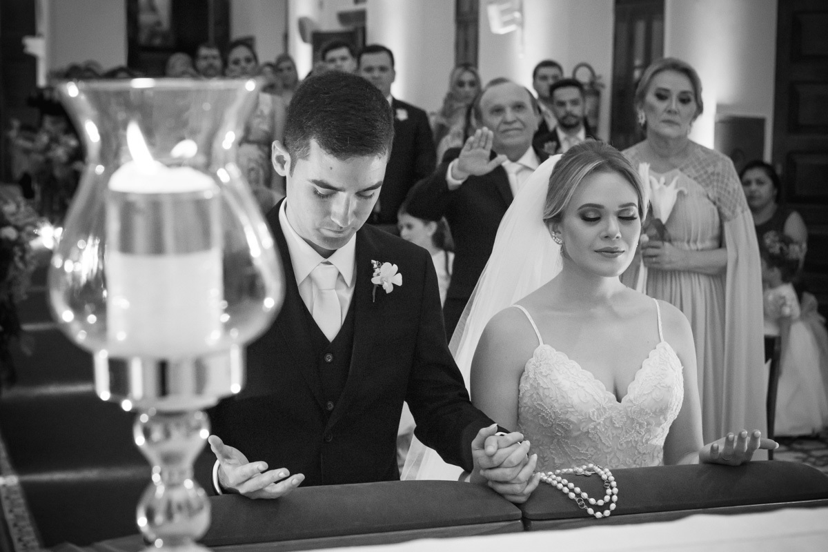 Celebração do casamento na Igreja Santa Terezinha, noivos ajoelhados de mãos dadas. e cabeça baixa e olhos fechados.
