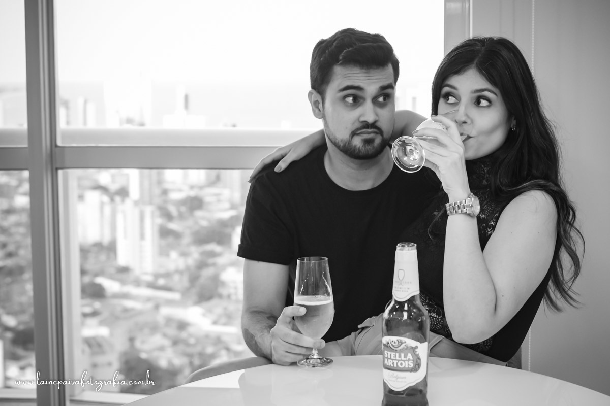 homem sentado na cadeira e namorada no colo, bebendo cerveja e ele olhando.