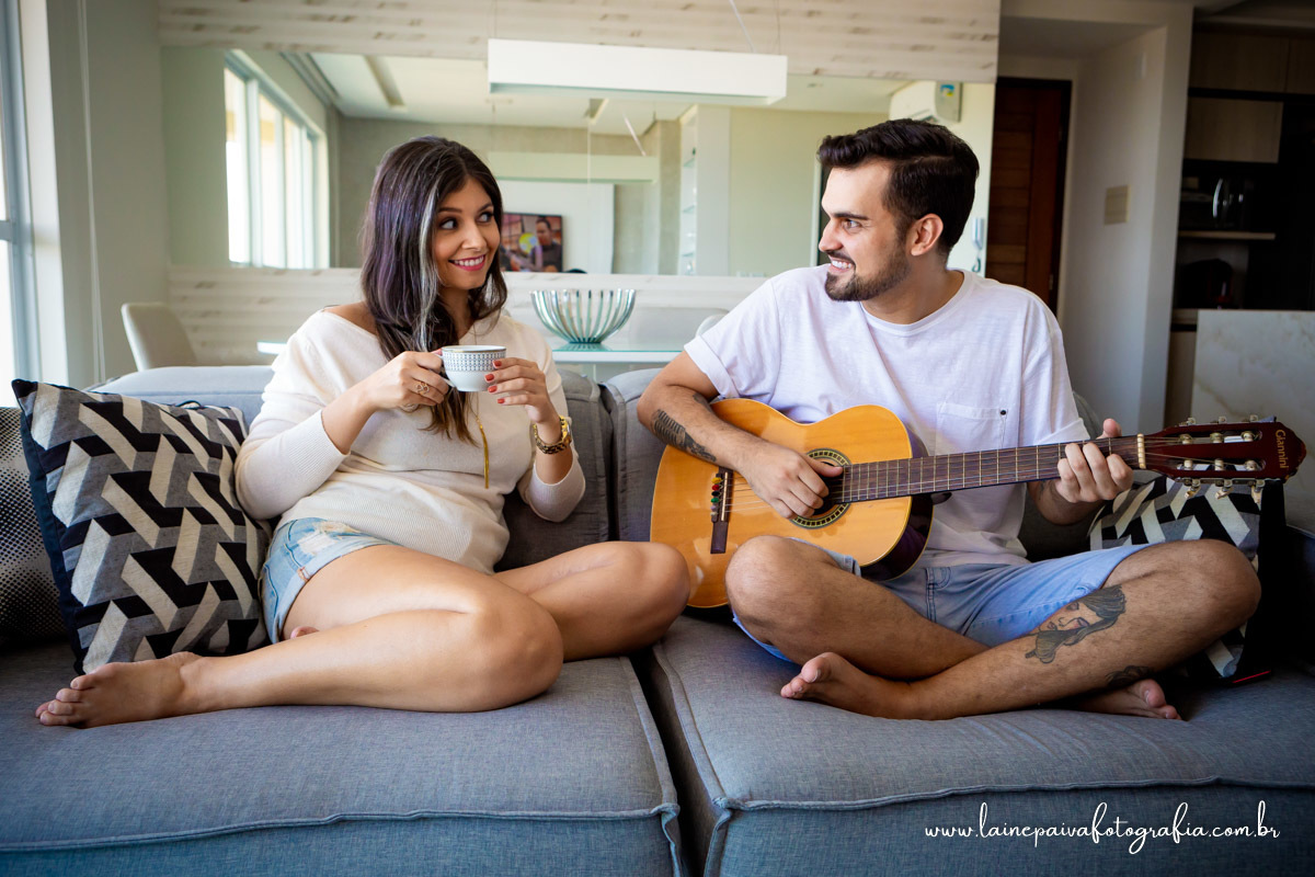 namorados sentados no sofá. Ela segurando uma xicara de cha na mão e ele tocando violão e se olhando.