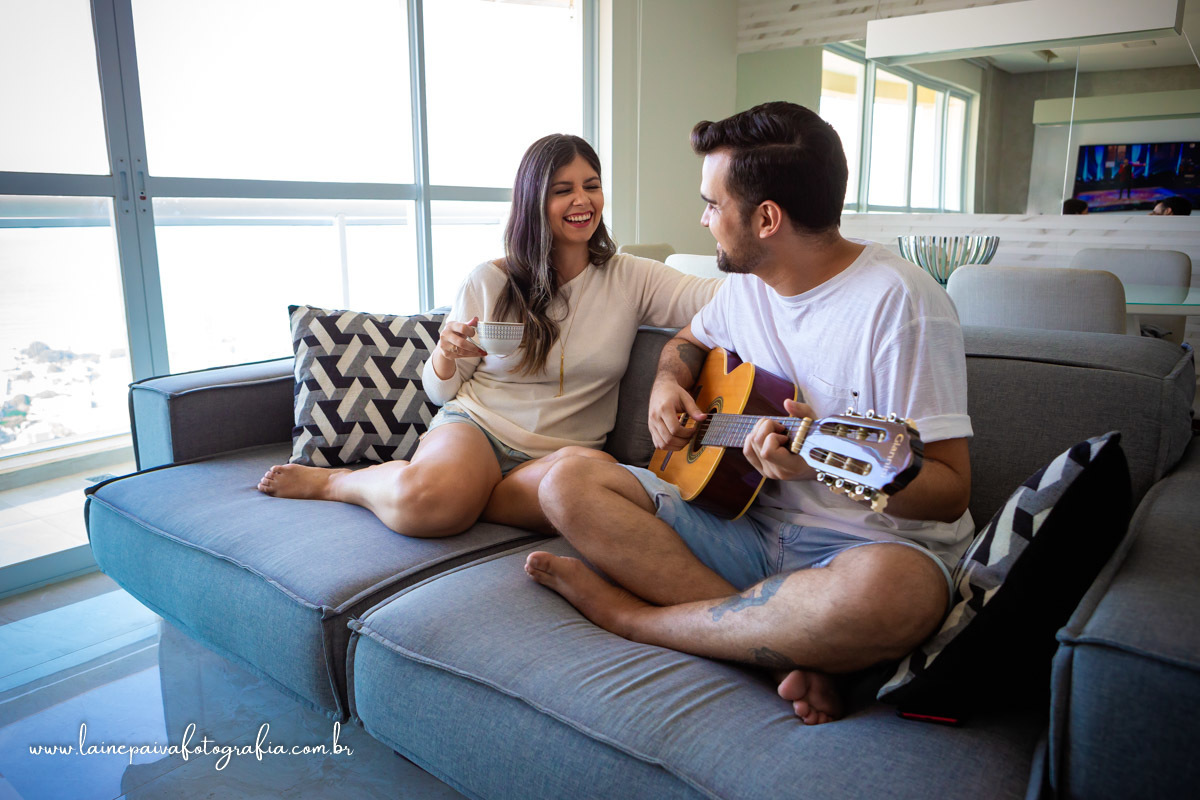 namorados sentados no sofá. Ela segurando uma xicara de cha na mão, sorrido e olhando para o namorado e ele tocando violão e se olhando.