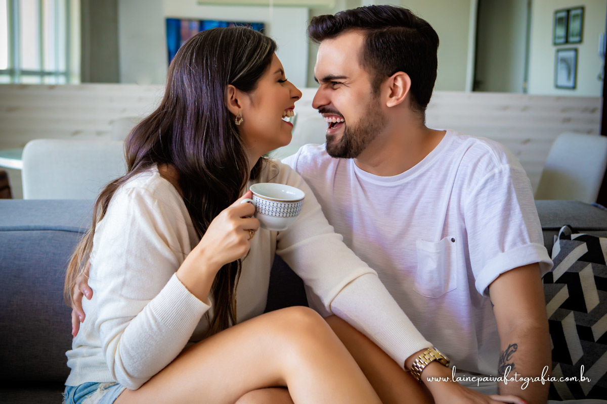namorados sentados no sofá. Ela segurando uma xicara de cha na mão, se olhando e rindo.