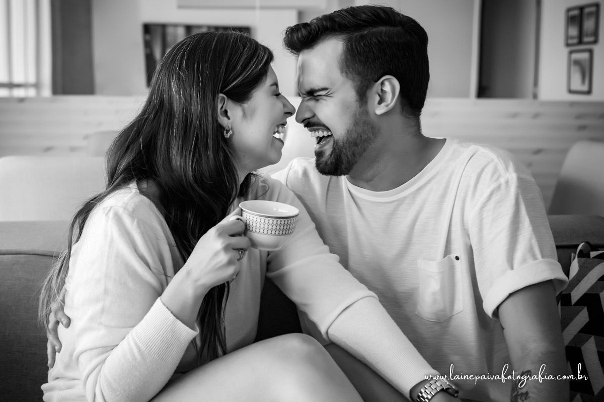 namorados sentados no sofá. Ela segurando uma xicara de cha na mão, se olhando e rindo com nariz encostado.
