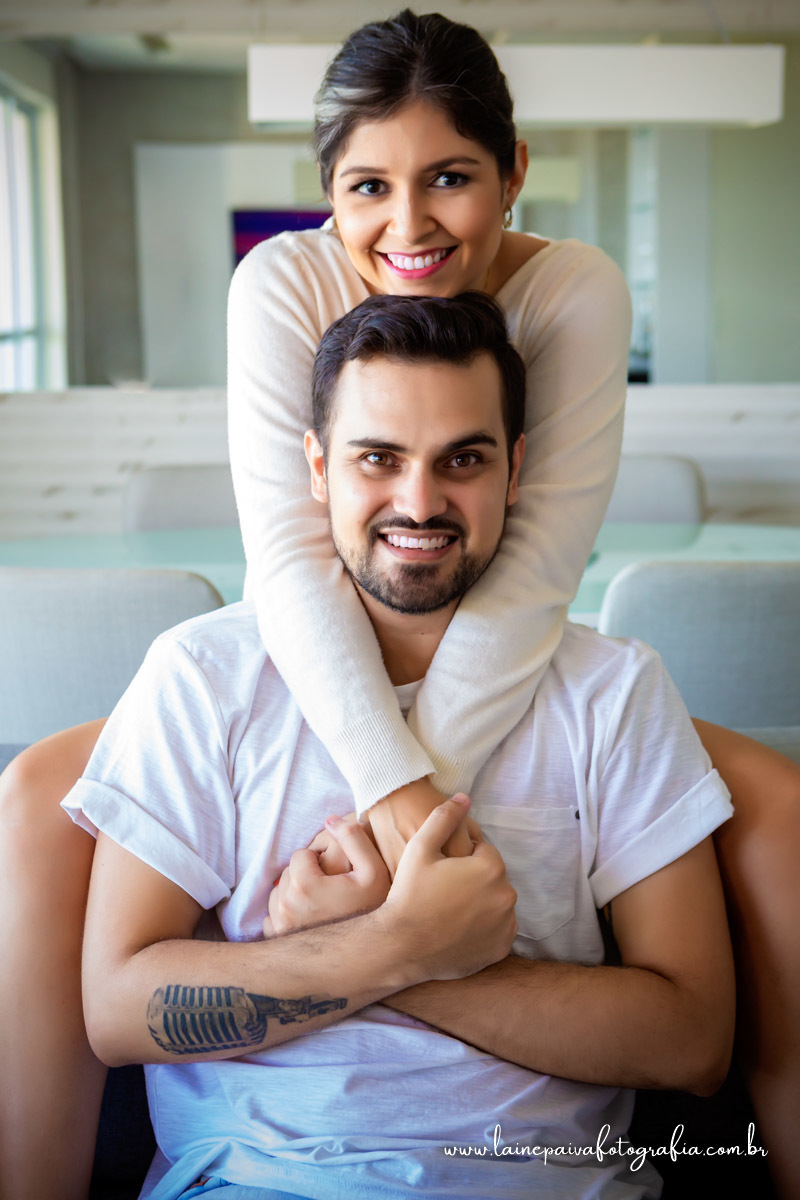 namorados sentados no sofá. Ela em cima abraçando ele e ambos olhando para a camera.