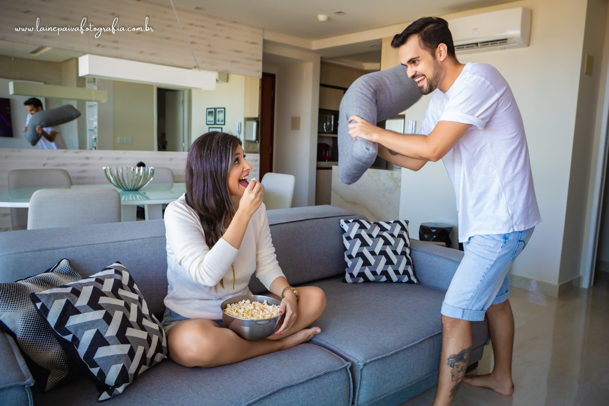 Namorada sentada no sofá comendo pipoca e namorado segurando almofada em pé para bater nela.