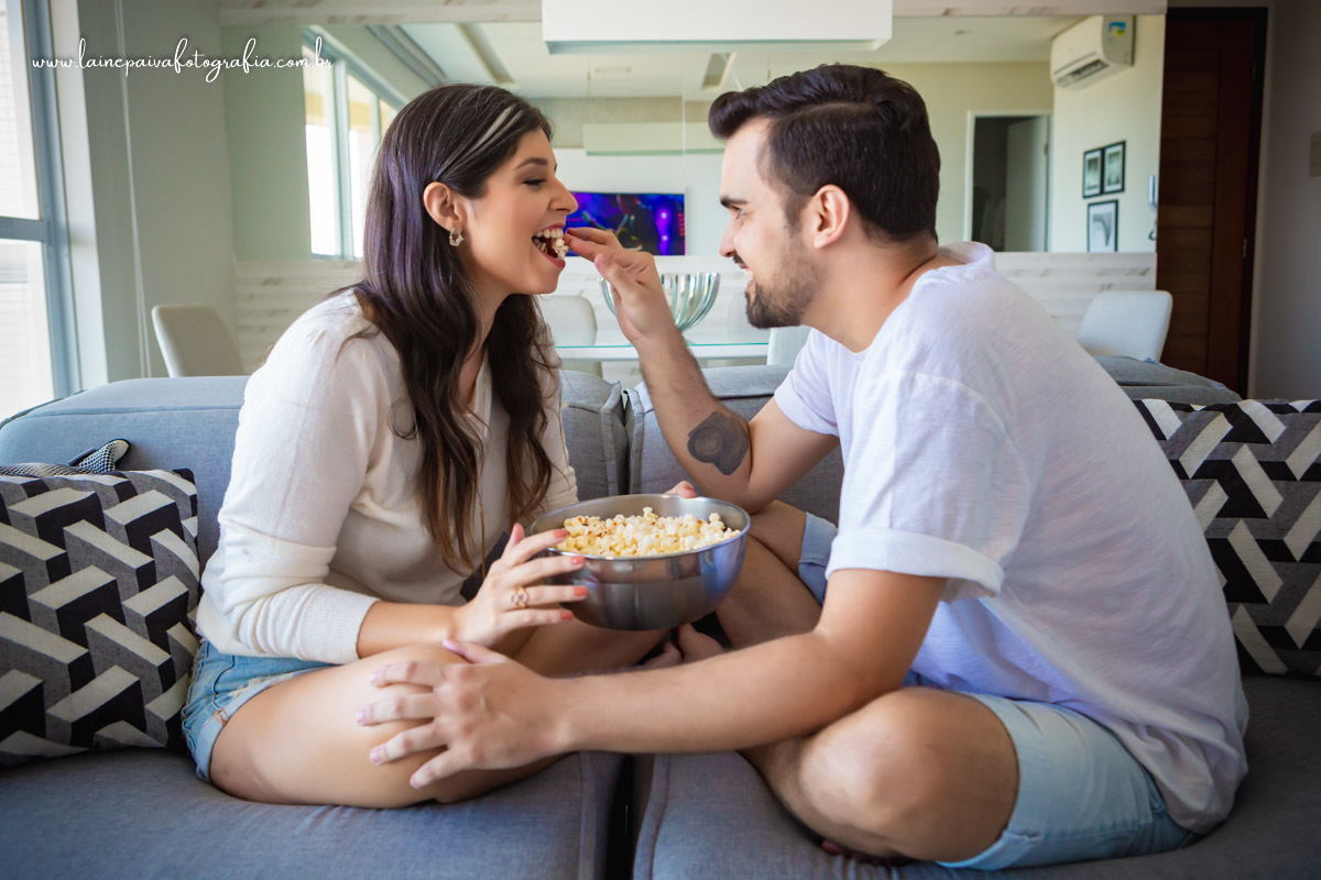 Namorados sentados no sofá com balde de pipoca, ele colocando pipoca na boca dela.