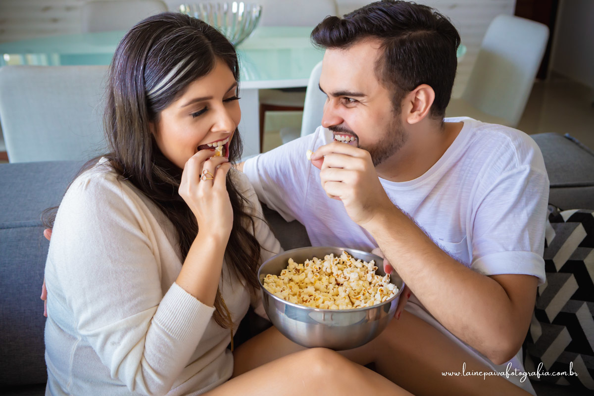Namorados sentados no sofá com balde de pipoca, ele colocando pipoca na boca dela.