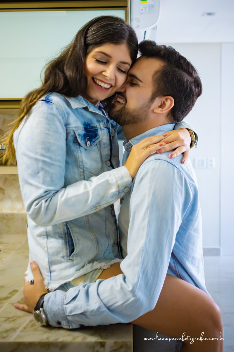 Namorada sentada em bancada abraçando namorado que esta em pé e beijando seu pescoço.
