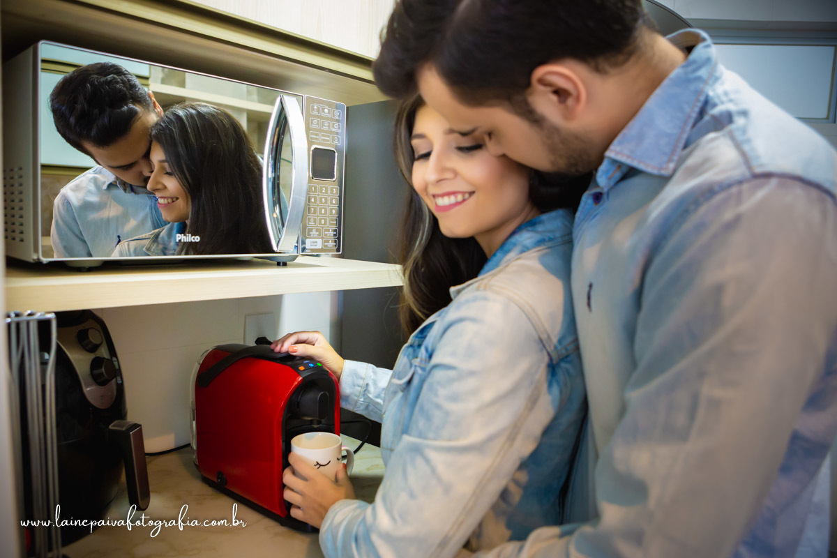 Casal em pé fazendo café, ele por trás dela abraçando-a e beijando-a com o reflexo no microondas.