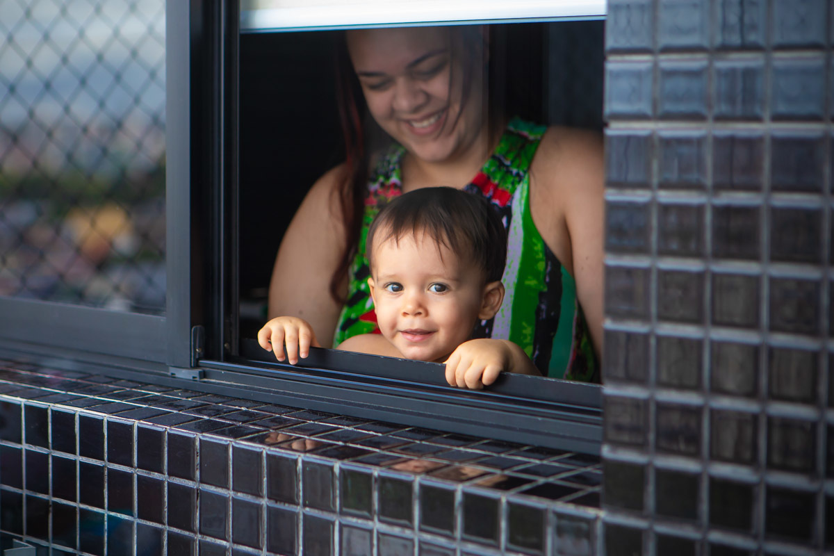 ensaio em familia em casa em salvador, criança em pé se segurando na janela e mãe atrás sorrindo.