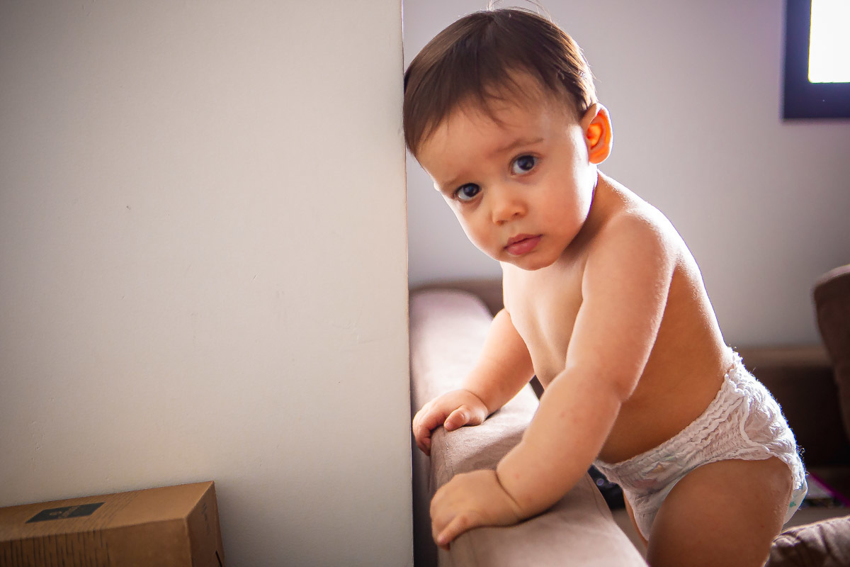 ensaio em familia em casa em salvador, menino em pé no sofá batendo a cabeça na parede e olhando