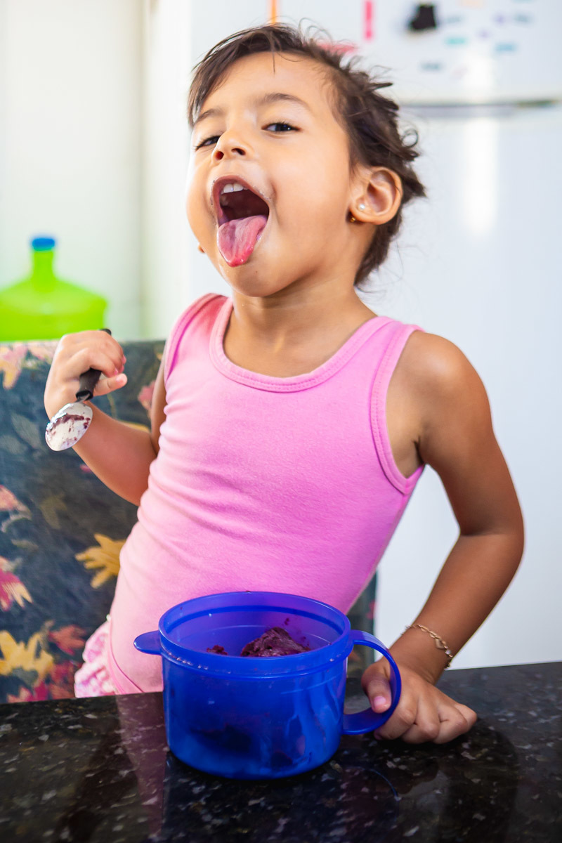 menina mostando a lingua melada de açai segurando uma colher na mão e em cima da cadeira.