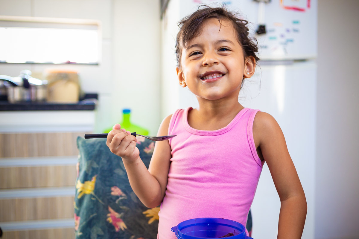 menina rindo com a boca melada de açai segurando uma colher na mão e em cima da cadeira.