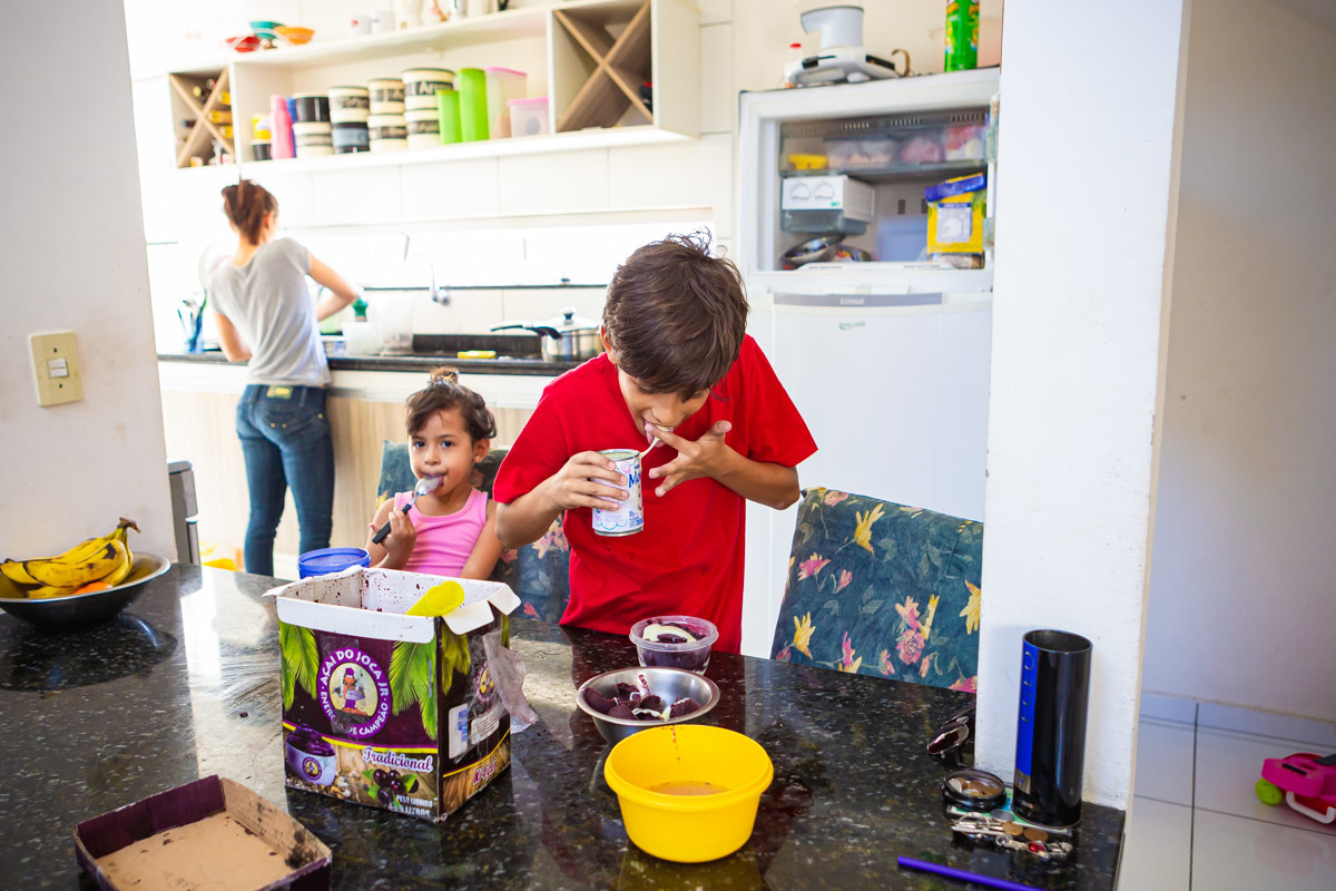 menino colocando na boca o dedo melado de leite condensado enquanto mãe lava a louça e irmã toma açai.