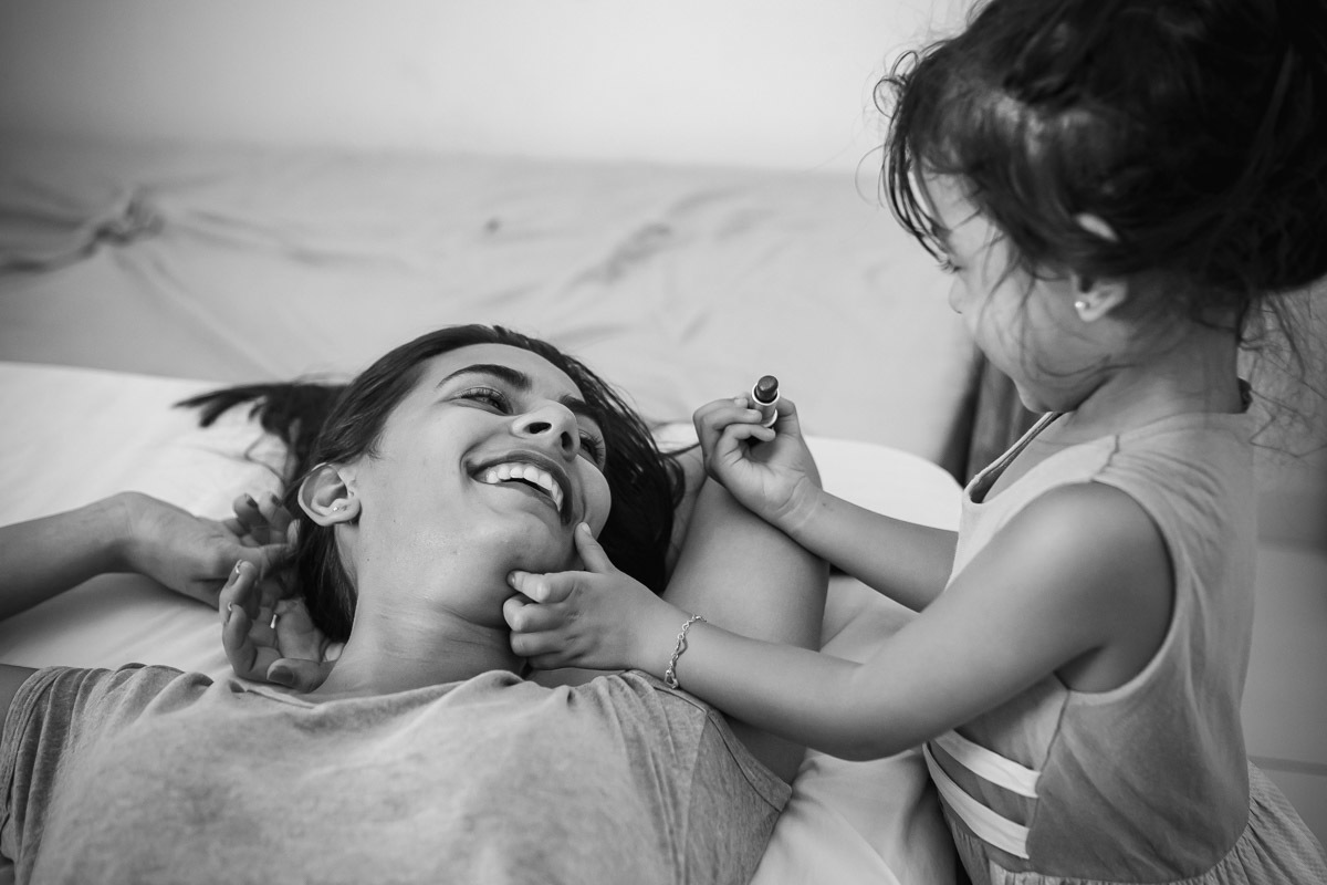 mãe deitada na cama e filha passando batom em sua boca.