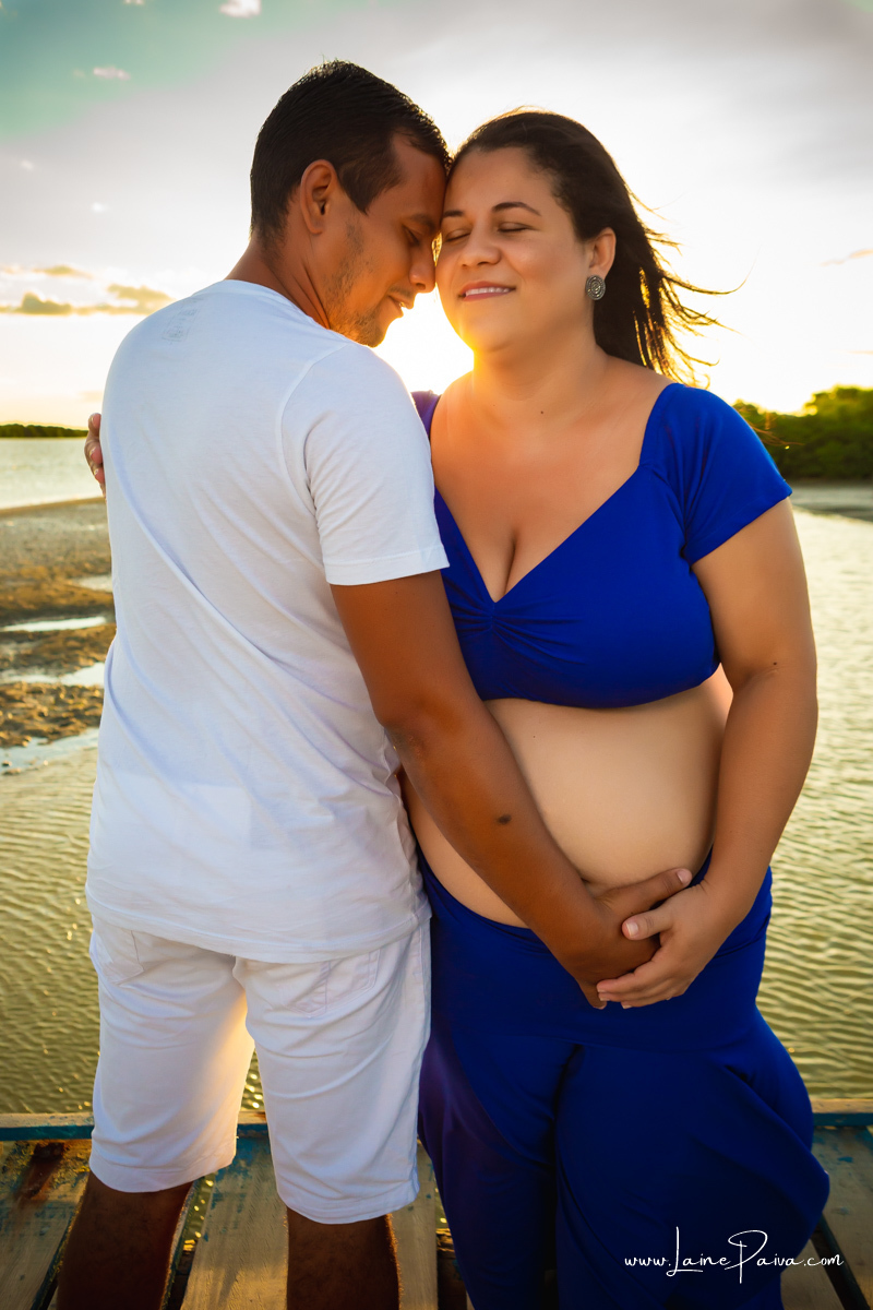 gestante na abraçada com o marido 
 com as cabeças encostadas e olhos fechados com mãos na barriga e sorrindo num ensaio no fim de  tarde num trapiche com um por do sol lindo.