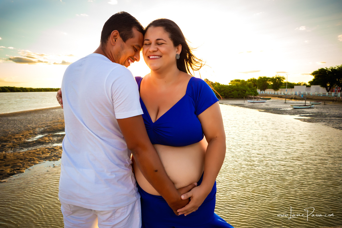 gestante na abraçada com o marido 
 com as cabeças encostadas e olhos fechados com mãos na barriga e sorrindo num ensaio no fim de  tarde num trapiche com um por do sol lindo.