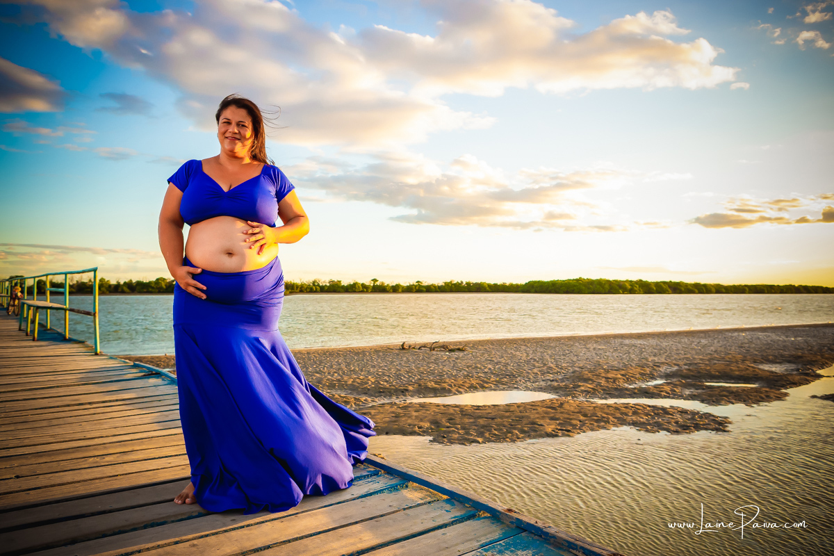 gestante usando saia e croped azul com mãos na barriga e sorrindo num ensaio no fim de  tarde num trapiche com um por do sol lindo.