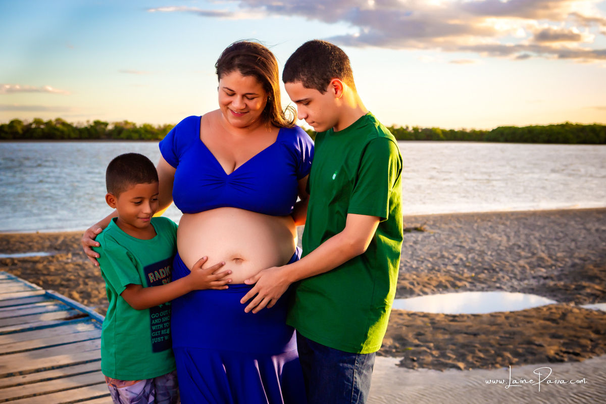 gestante usando saia e croped azul, com filho e enteado com mãos na barriga e sorrindo num ensaio no fim de  tarde num trapiche com um por do sol lindo.