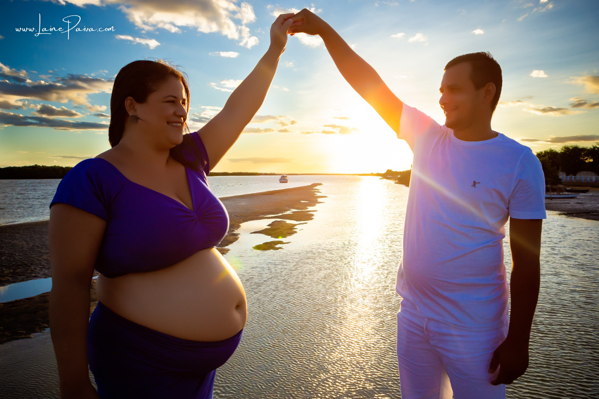 gestante com marido segurando sua mão para cima em ensaio no fim de  tarde num trapiche com um por do sol lindo e passaros voando.