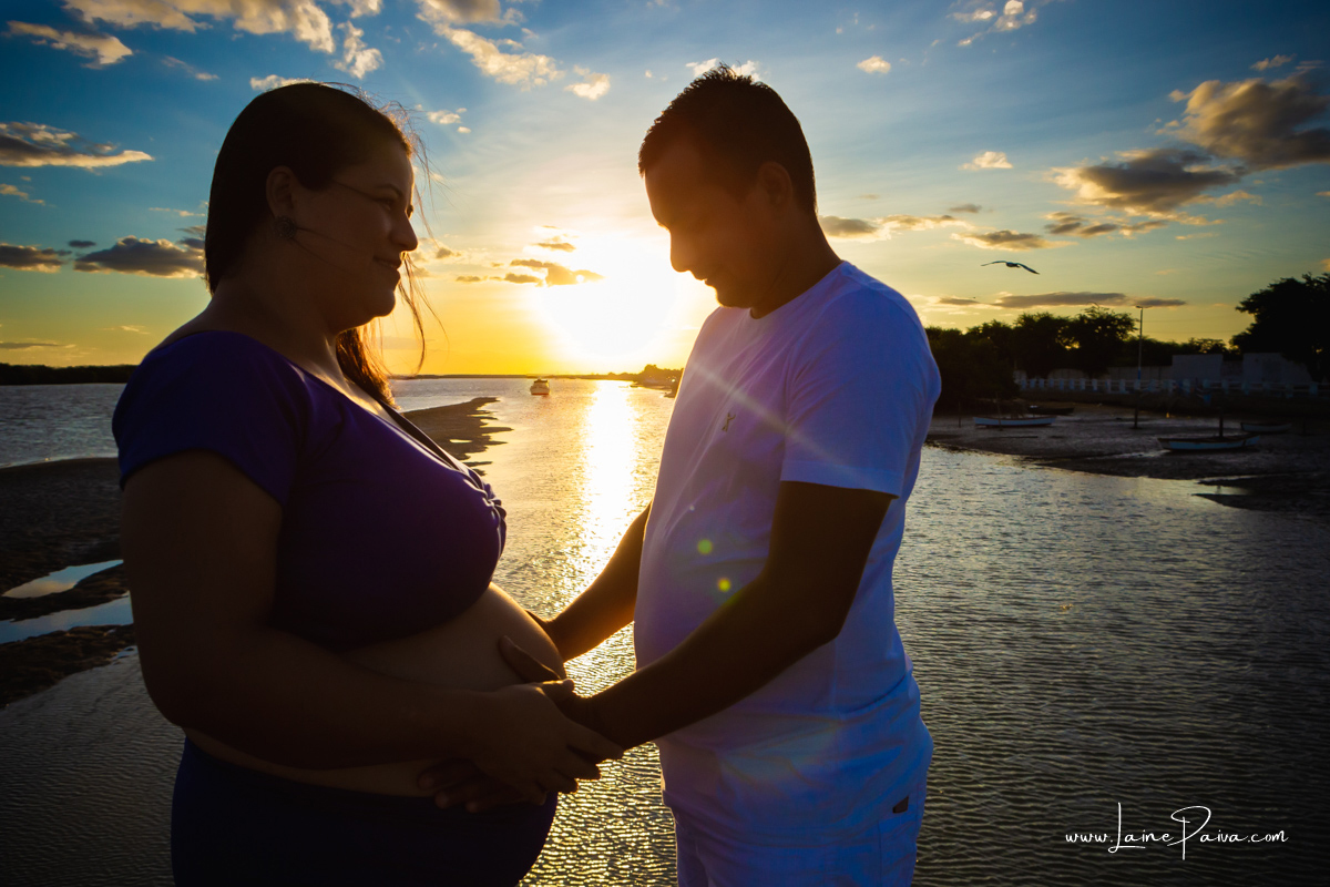 gestante com marido de frente para o outro segurando na barriga ensaio no fim de  tarde num trapiche com um por do sol lindo e passaros voando.