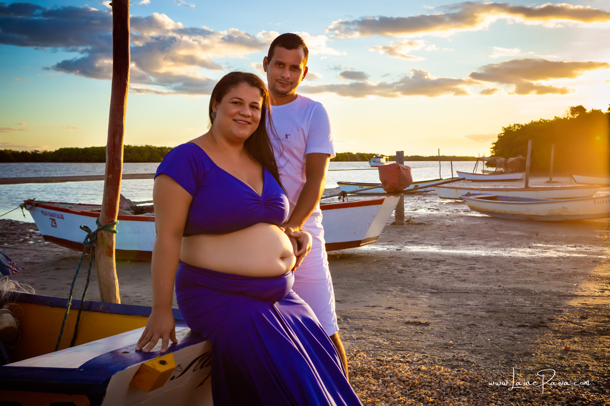 gestante sentada em barco na beira do rio com o marido em pé ao seu lado com mão na barriga num ensaio no fim de  tarde num trapiche com um por do sol lindo.