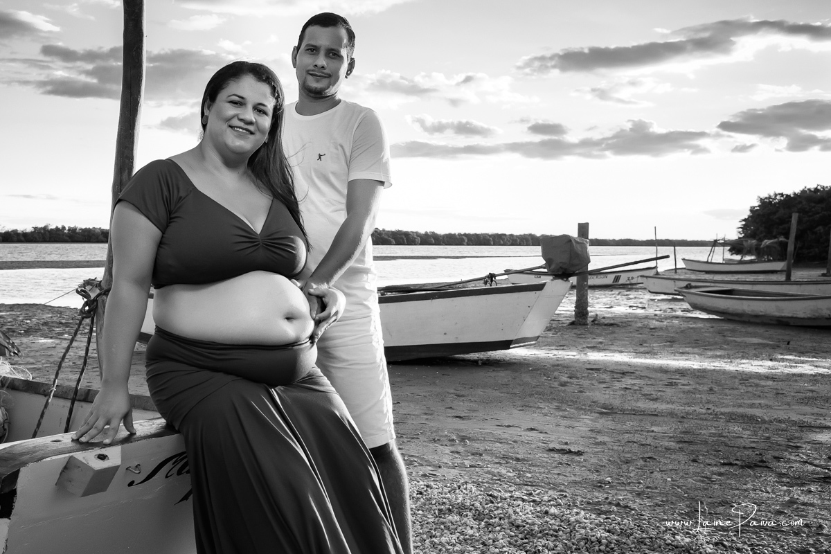 gestante sentada em barco na beira do rio com o marido em pé ao seu lado com mão na barriga num ensaio no fim de  tarde num trapiche com um por do sol lindo.