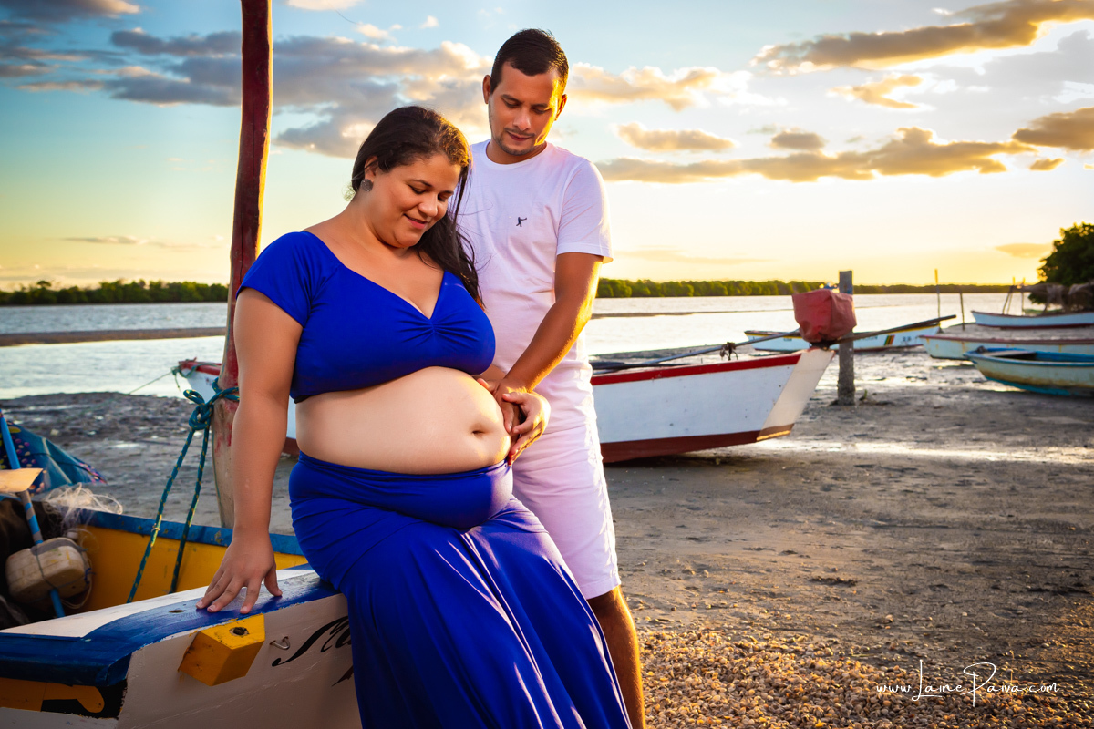 gestante sentada em barco na beira do rio com o marido em pé ao seu lado com mão na barriga e olhando para ela num ensaio no fim de  tarde num trapiche com um por do sol lindo.