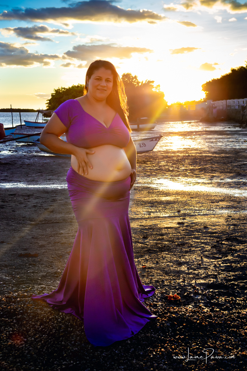 gestante em pé usando roupa azul com mão na barriga no meio de barcos proximo ao rio num ensaio no fim de  tarde num trapiche com um por do sol lindo.
