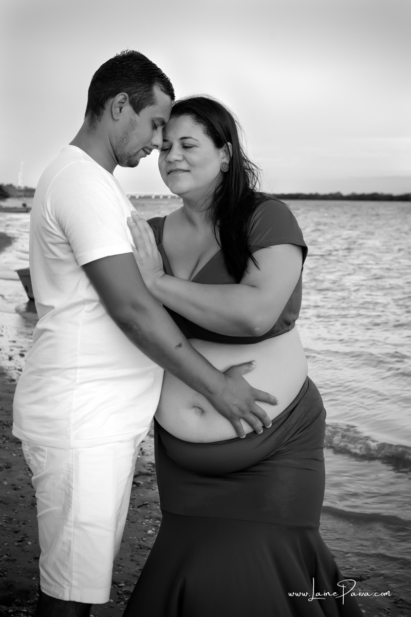 gestante abraçada a marido na beira do rio com cabeça coladas, olhos fechados e com as mãos na barriga num ensaio no fim de  tarde num trapiche com um por do sol lindo.