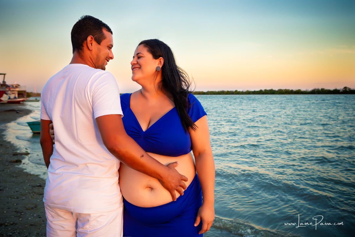 gestante abraçada a marido na beira do rio com se olhando, sorrindo e com as mãos na barriga num ensaio no fim de  tarde num trapiche com um por do sol lindo.