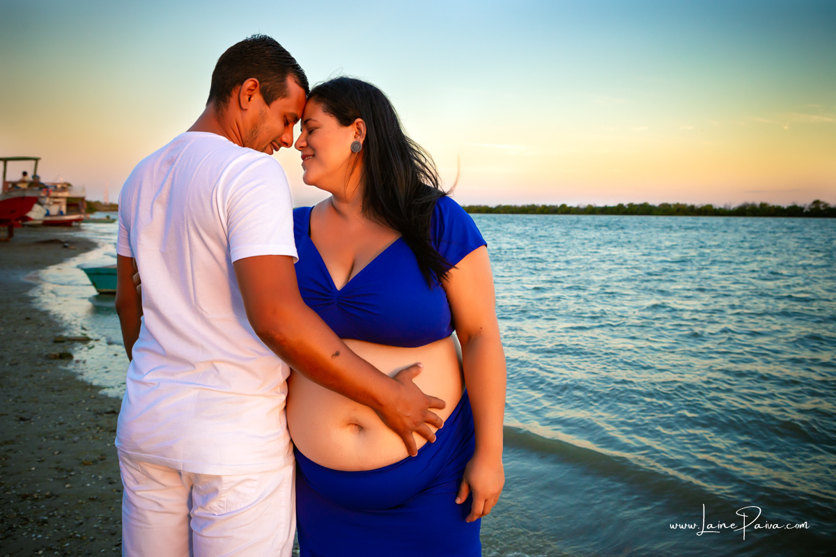 gestante abraçada a marido na beira do rio com cabeça coladas, olhos fechados, sorrindo e com as mãos na barriga num ensaio no fim de  tarde num trapiche com um por do sol lindo.