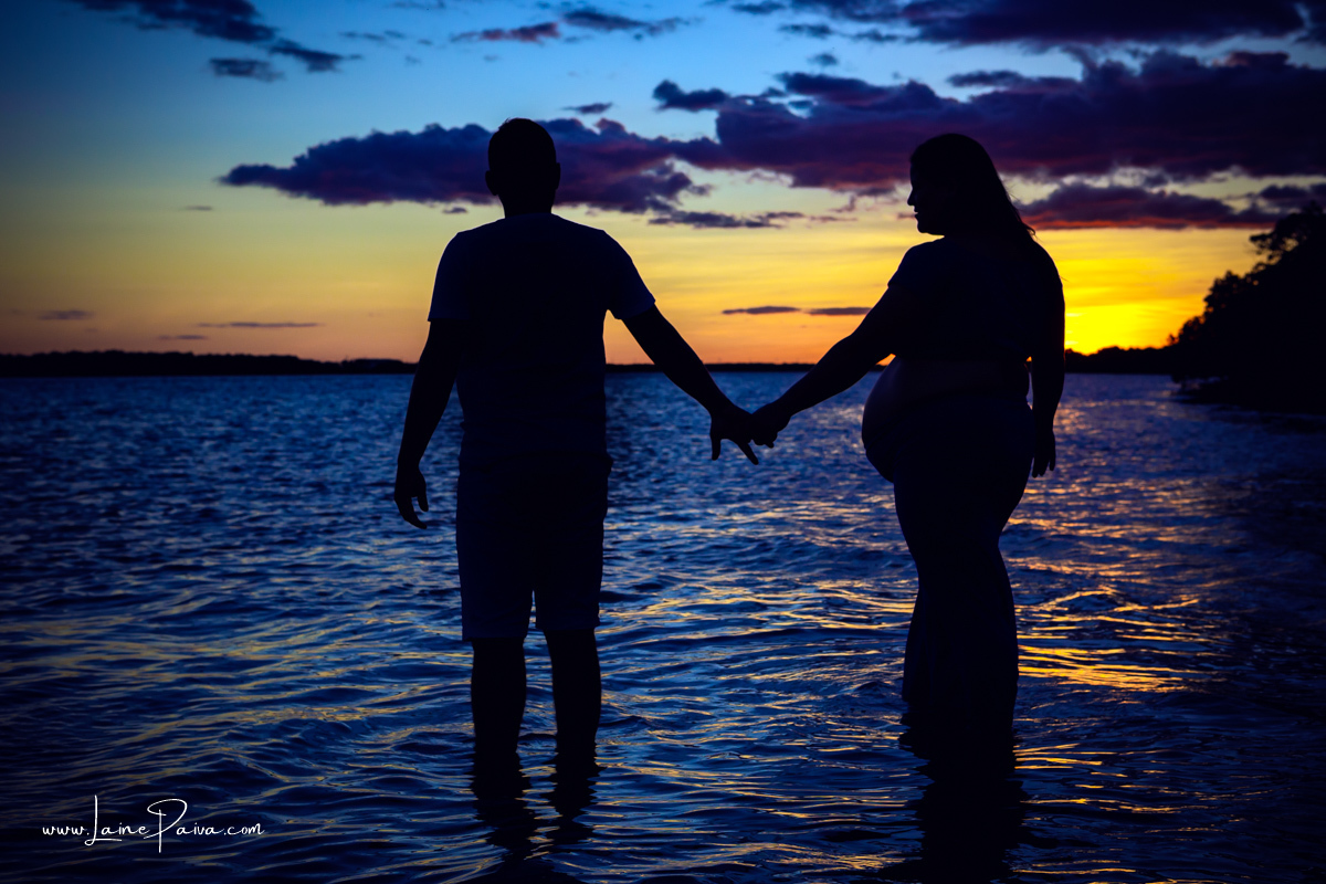silhueta de gestante caminhando com marido dentro de um rio  de mãos dadas num ensaio no fim de  tarde num trapiche com um por do sol lindo.