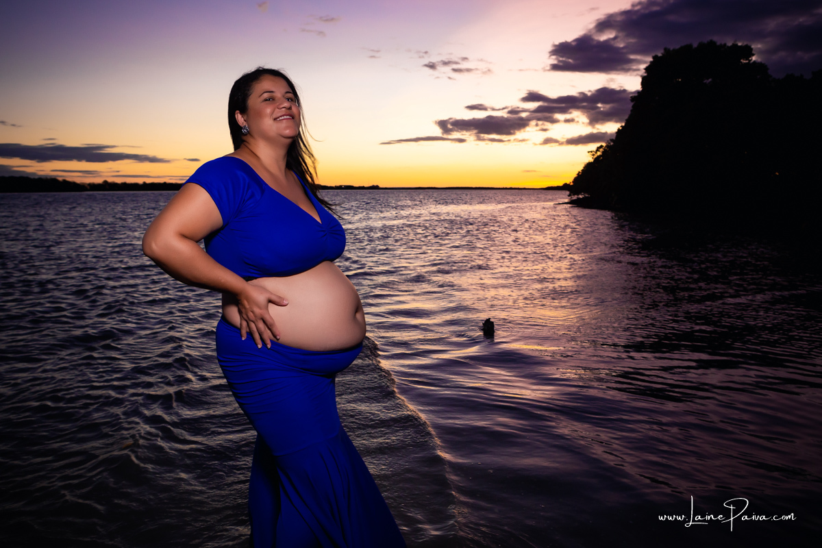 gravida dentro do rio com roupa azul e mãos na barriga sorrindo num trapiche com um por do sol lindo.