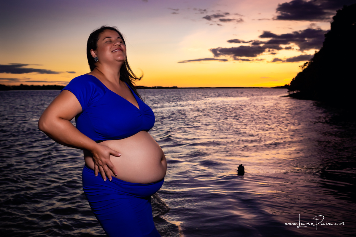 gestante dentro do rio com roupa azul e mãos na barriga sorrindo num trapiche com um por do sol lindo.