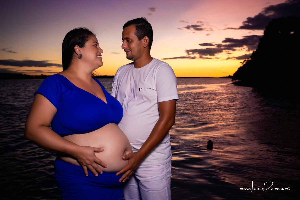 gravida e seu marido dentro do rio com mãos na barriga sorrindo num trapiche com um por do sol lindo.