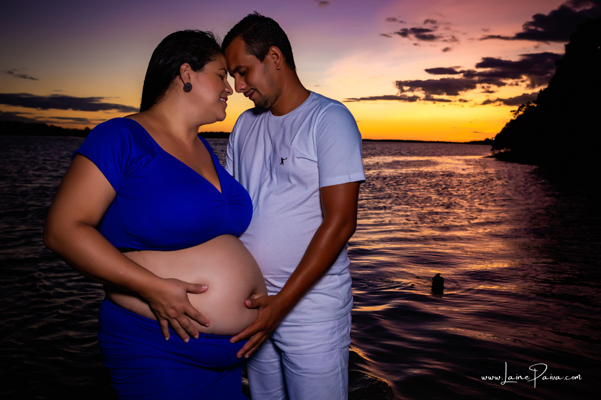 gravida e seu marido dentro do rio com mãos na barriga, cabeças coladas e sorrindo num trapiche com um por do sol lindo.