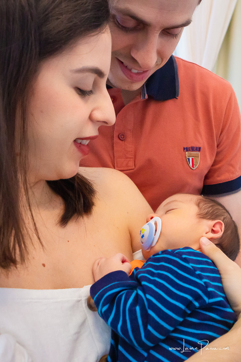 bebe dormindo no colo da mãe que esta abraçada com marido e olhando para o filho em ensaio lifestyle de newborn em casa