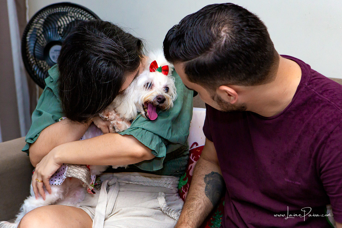 casal sentado em sofá da casa com cachorra no colo da mulher recebendo cheiro em ensaio lifestyle em casa com a familia em natal 