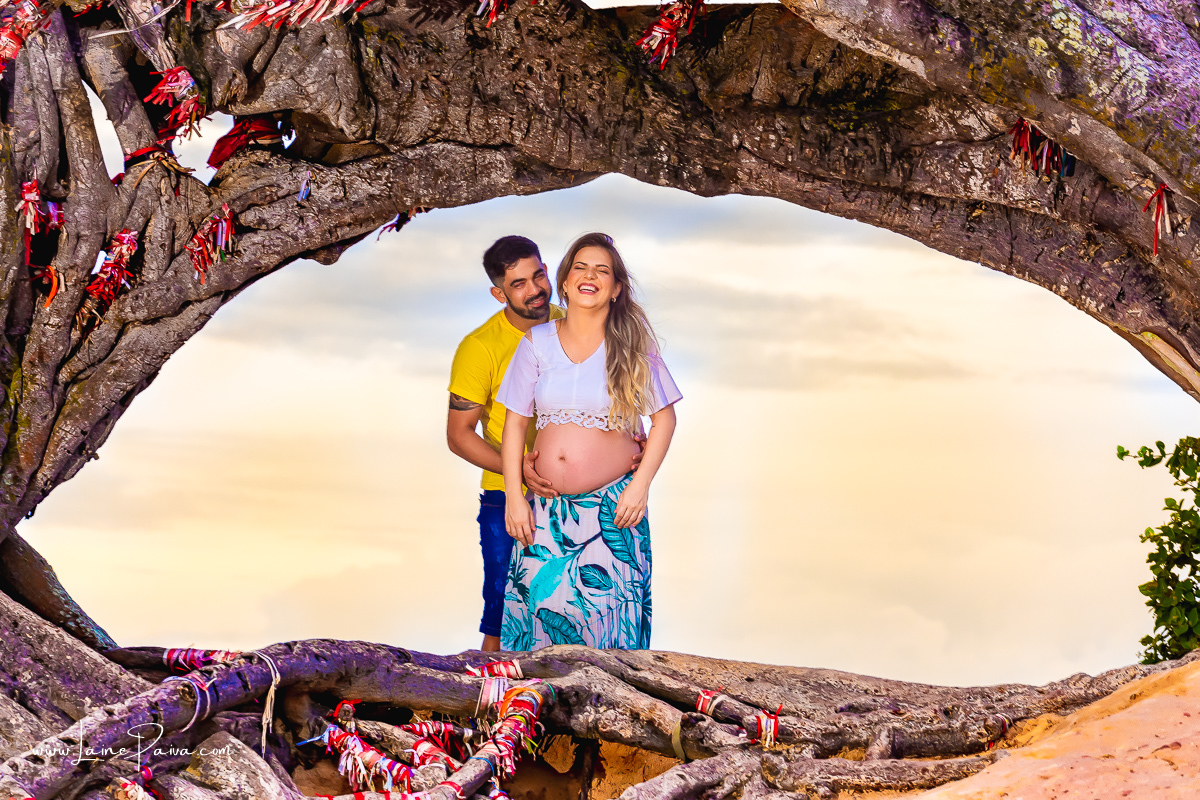 gestante sendo abraçada pelo marido por trás e sorrindo após ele falar algo em seu ouvido, dentro de tronco da árvore no ensaio gestante externo na praia na Árvore do amor.