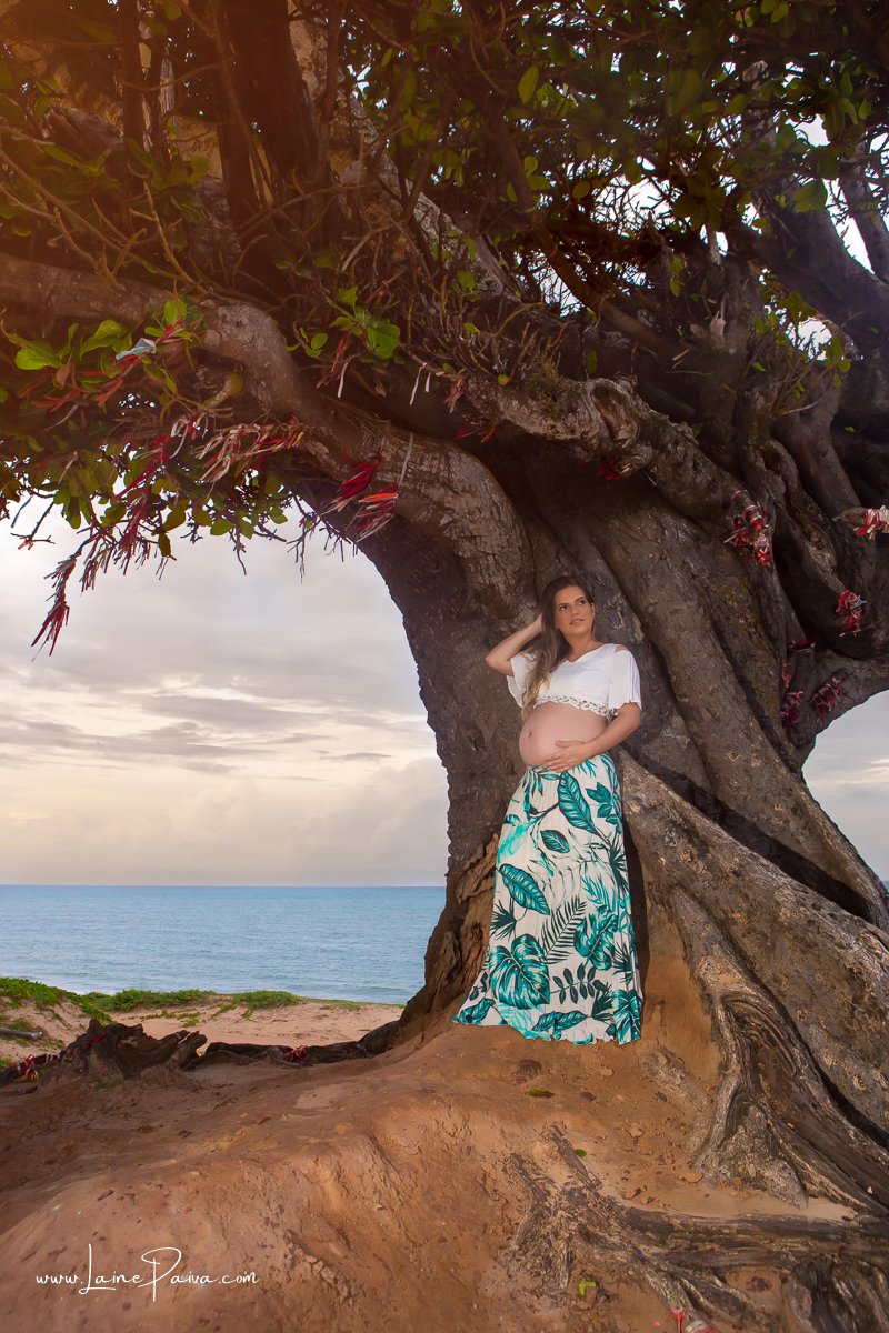 gravida com uma mão no cabelo e outra na barriga e encostada em árvore no ensaio gestante externo na praia na Árvore do amor.