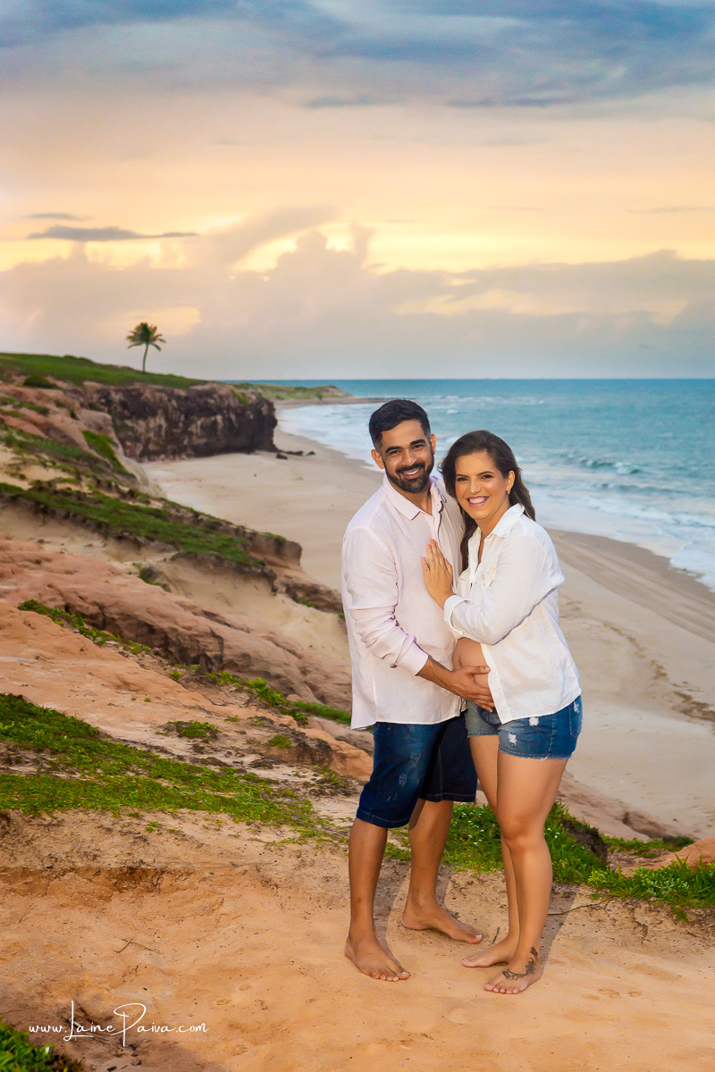 gestante com mão no peito do marido que tem a mão em sua barriga e sorrindo no ensaio gestante externo na praia na arvore do amor.