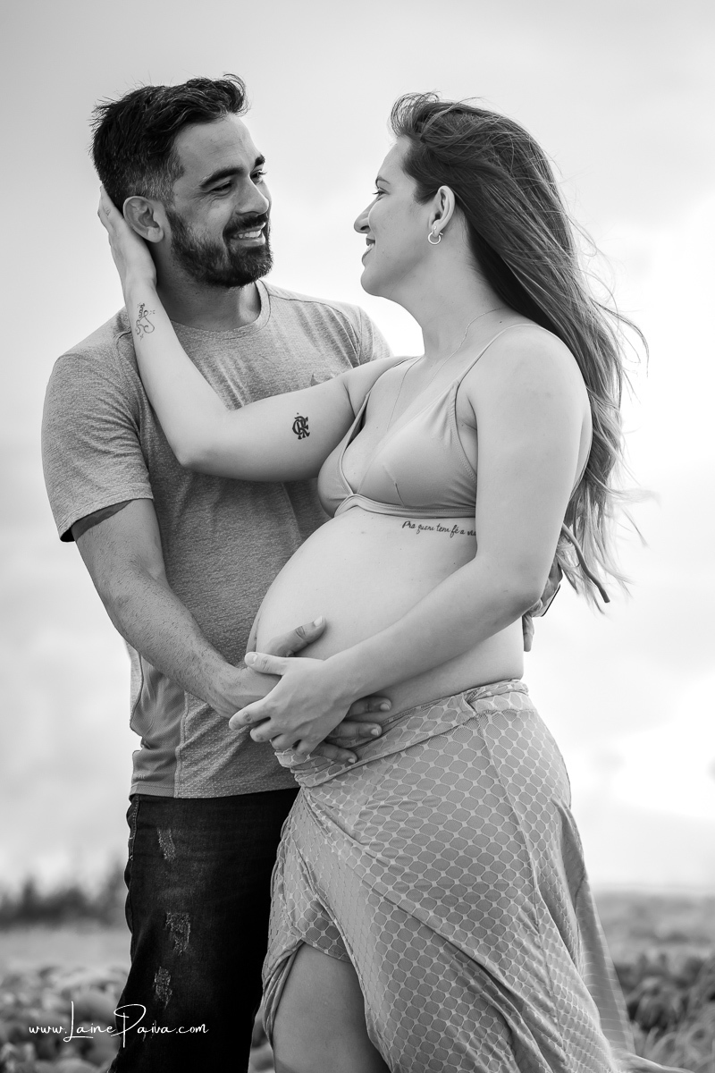 gestante com mão no rosto do marido e o olha sorrindo, marido com a mão em sua barriga no ensaio gestante externo na praia.