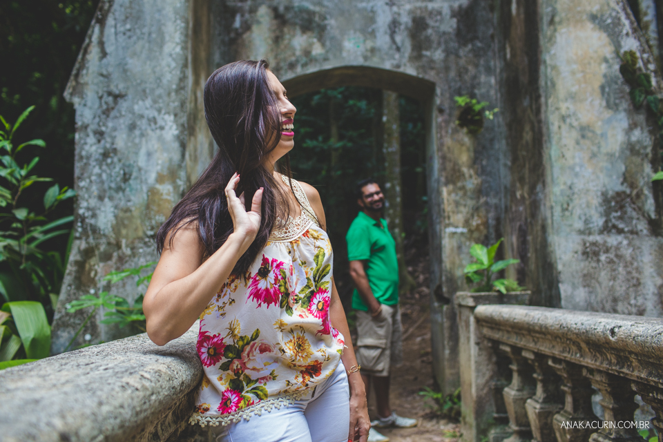 Mulher sorrindo espontaneamente  no meio de uma bonita ponte de pedra, enquanto seu noivo a admira do  portal. Fotografia de casal feita por Ana Kacurin no Rio de Janeiro - RJ.