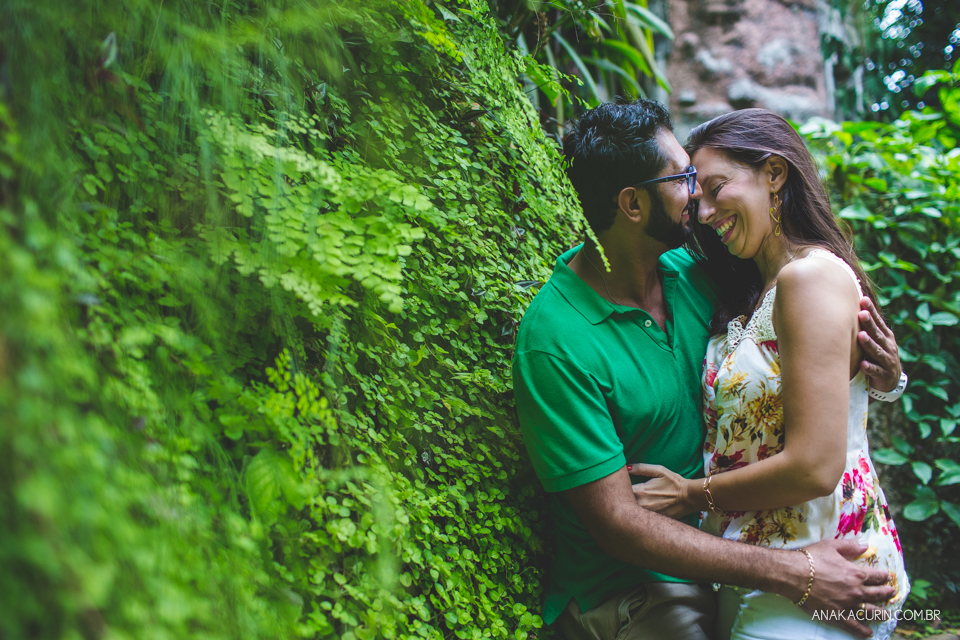 Casal se abraçando no meio das folhagens. Ao fundo, há uma torre antiga de pedra. Fotografia de casal feita por Ana Kacurin no Rio de Janeiro - RJ.