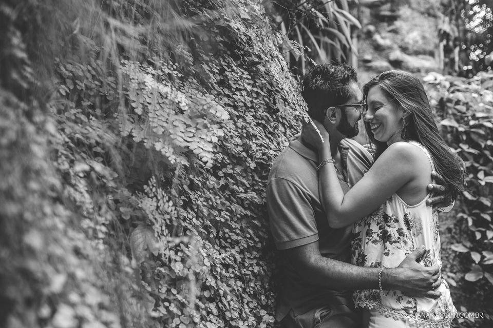 Casal se abraçando no meio das folhagens. Ao fundo, há uma torre antiga de pedra. Fotografia de casal feita por Ana Kacurin no Rio de Janeiro - RJ.