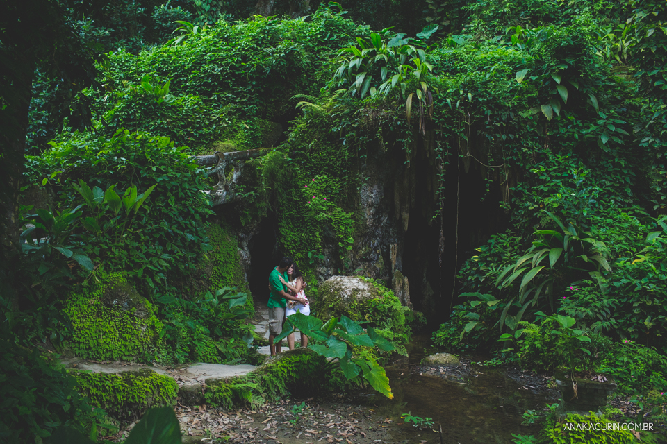Casal se abraçando no meio das folhagens. Ao fundo, há uma caverna. Fotografia de casal feita por Ana Kacurin no Rio de Janeiro - RJ.