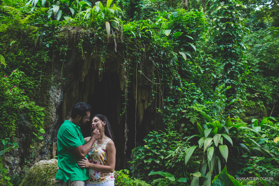 Casal se abraçando no meio das folhagens. Ao fundo, há uma caverna. Fotografia de casal feita por Ana Kacurin no Rio de Janeiro - RJ.