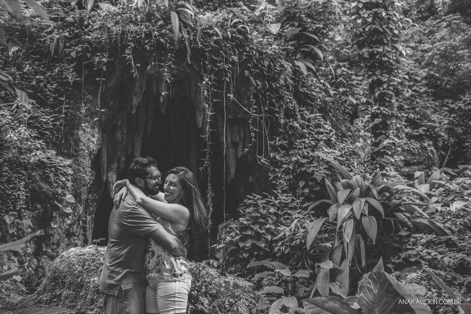 Casal se abraçando no meio das folhagens. Ao fundo, há uma caverna. Fotografia de casal feita por Ana Kacurin no Rio de Janeiro - RJ.